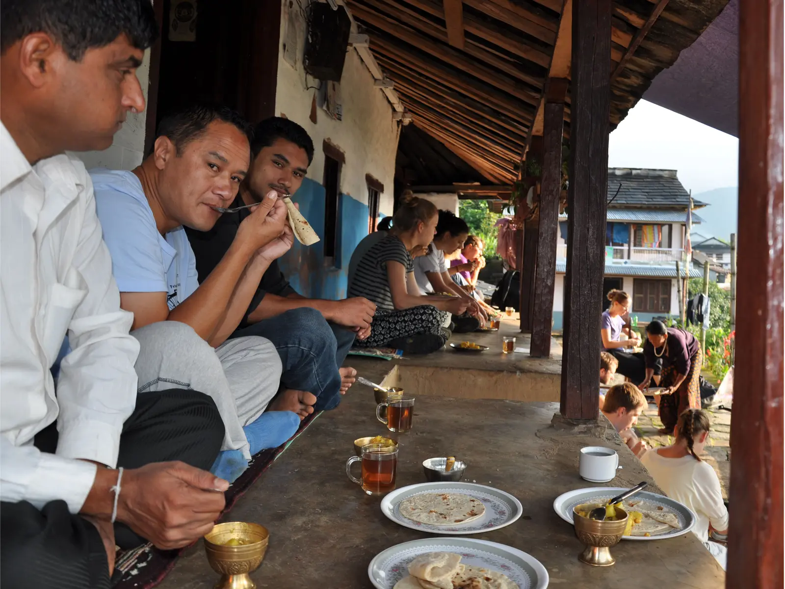 Hosts and visitors eating roti and curry together on a long veranda