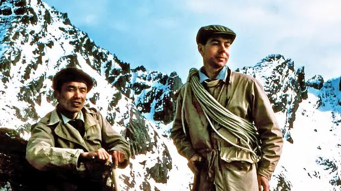 Two men in caps and coats, one carrying a coiled rope, in front of snowy peaks