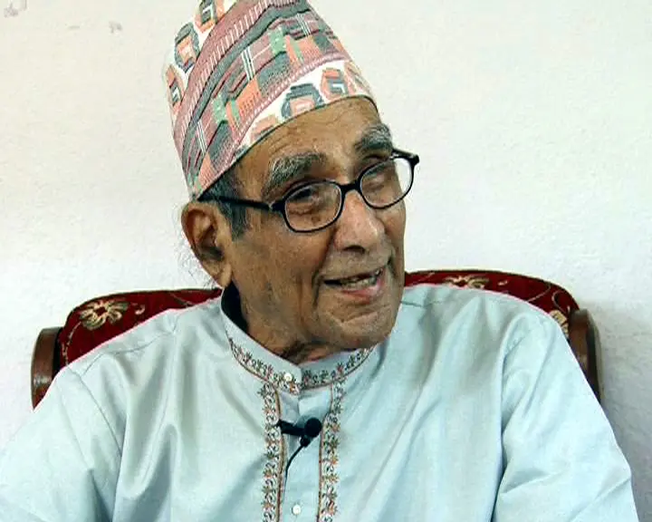 Elderly man in a dhaka topi and glasses speaking while seated