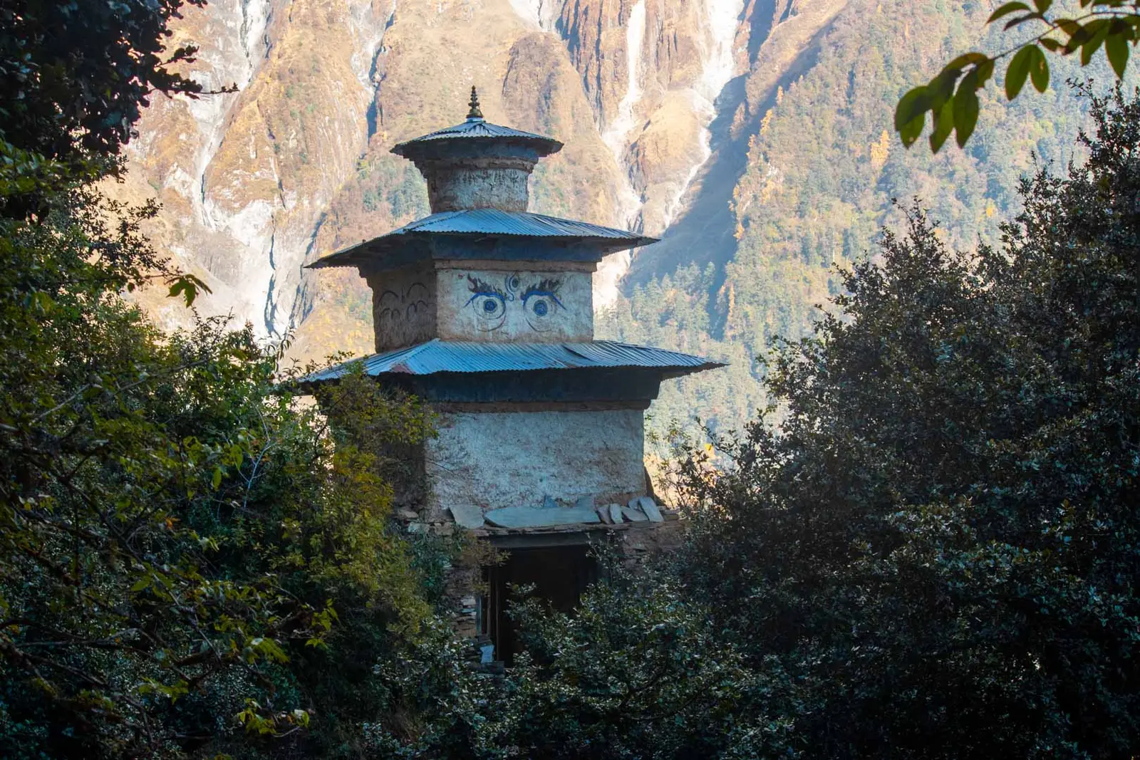Stone chorten with painted eyes and tiered metal roofs among trees