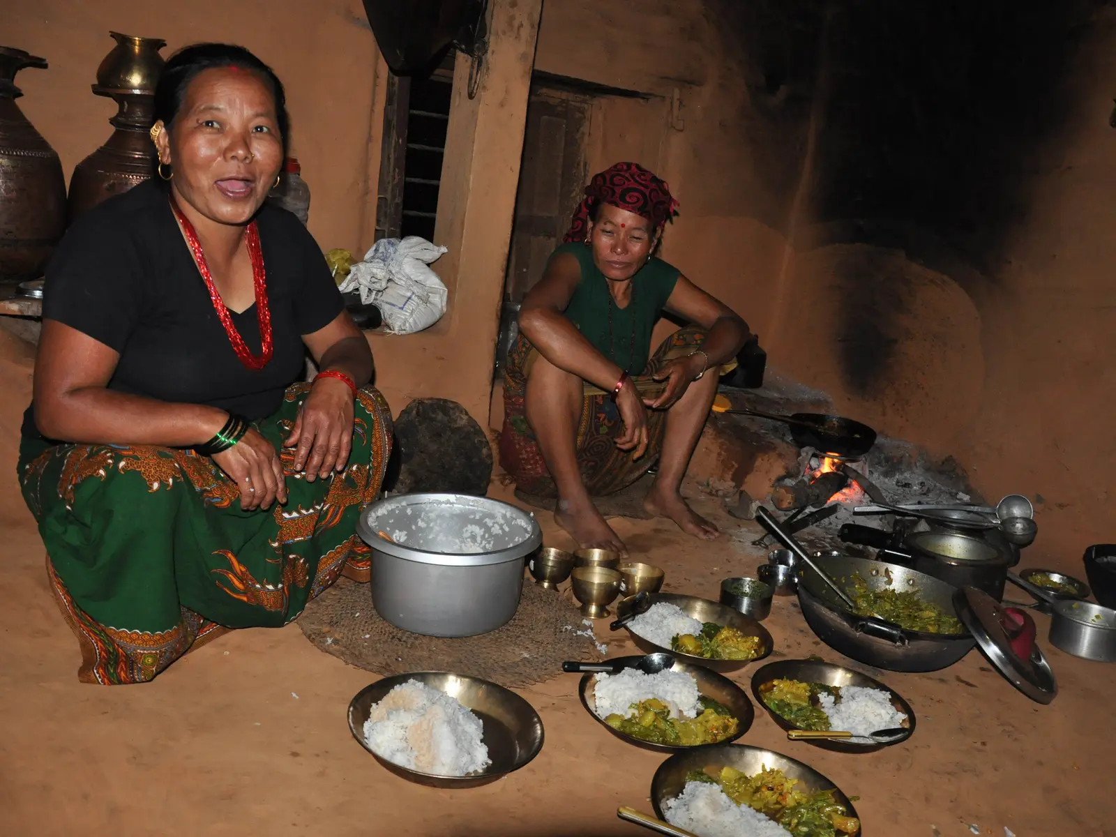 Two women serving rice and curry beside a wood fire in a village kitchen