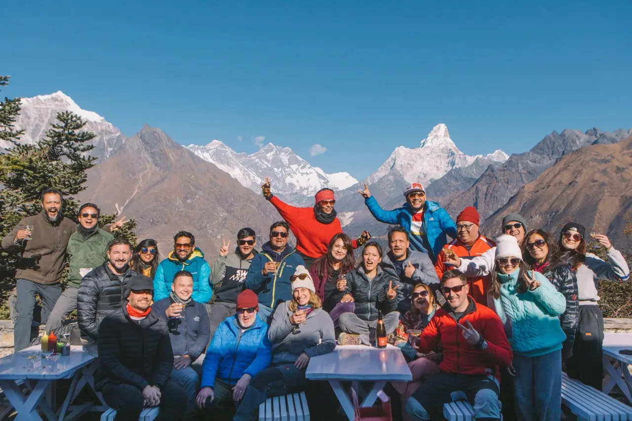 A group of people cheering with drinks on a terrace, with snow-capped Himalayan peaks behind them.