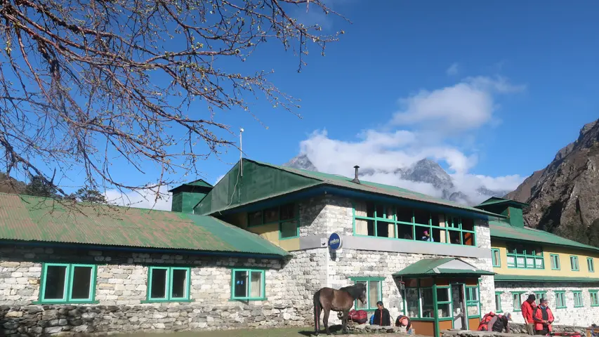 Stone lodge with a green roof, with a horse and trekkers outside below a snowy peak